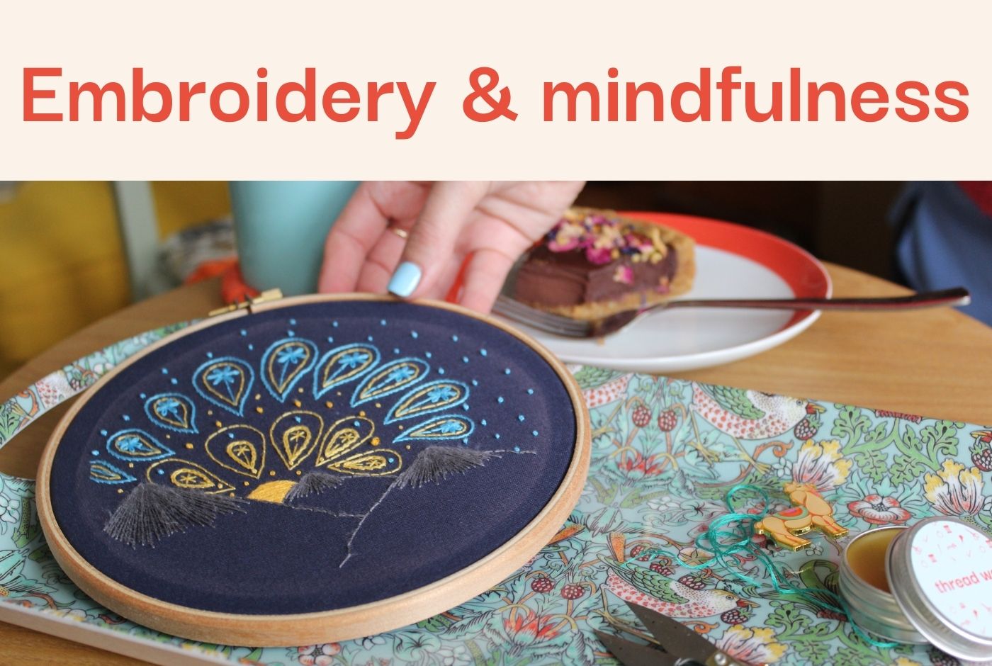 A hand holds embroidery with a sunrise design on navy fabric on a patterned tray with a slice of cake in the background.
