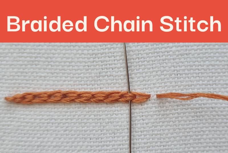 A close up of Braided Chain Stitch embroidered in orange thread on white fabric, with a needle sticking through the thread.