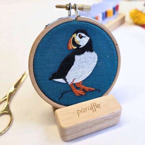 Embroidery hoop standing on an embroidery hoop stand, with puffin design on a blue background, branded 'paraffle'.