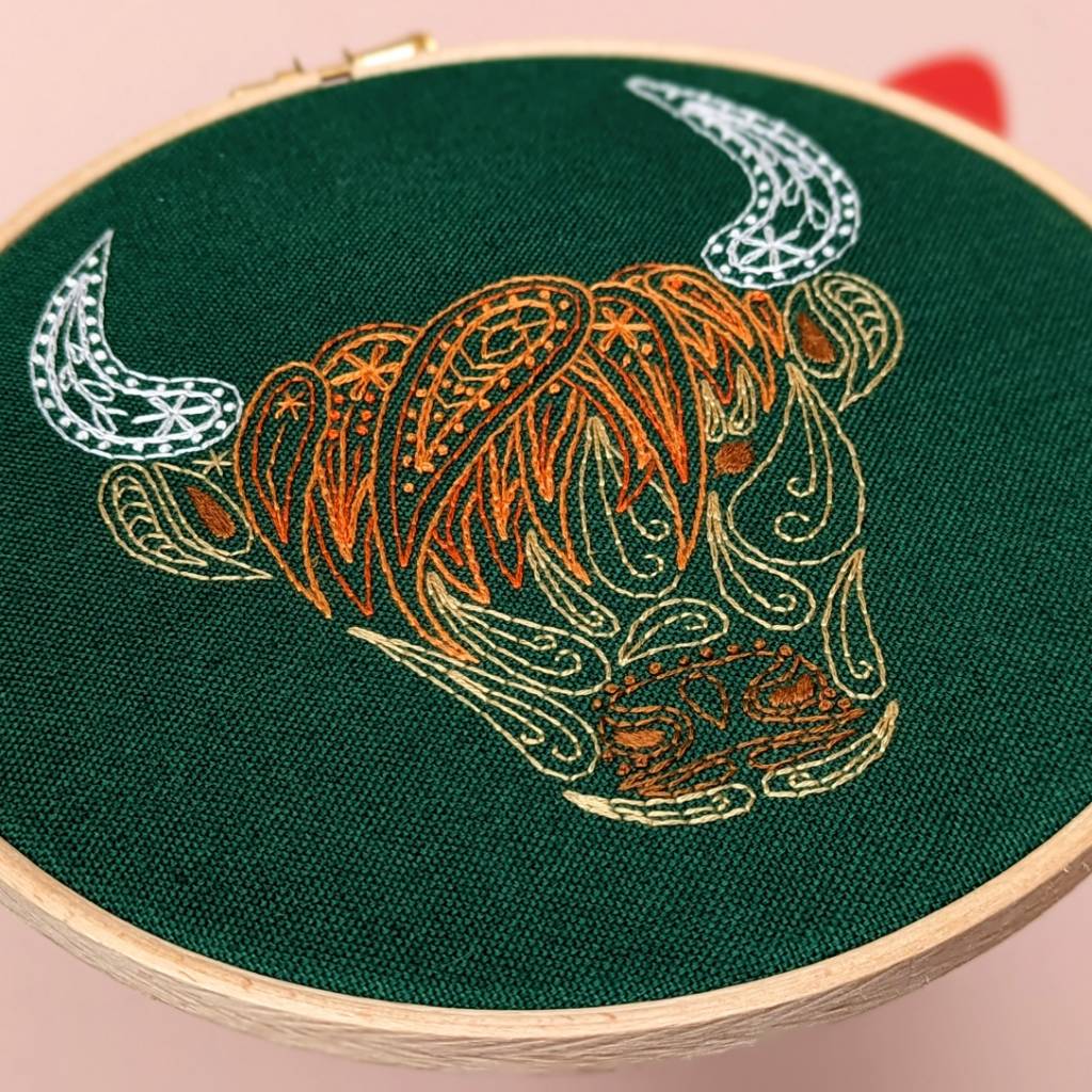 Angled photo of a orange &amp; brown Highland Cow embroidered in a paisley style on green fabric