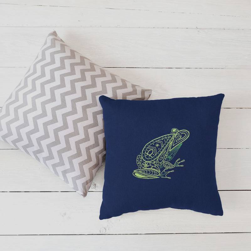 A navy blue cushion embroidered with a paisley style green frog design rests on white floorboards and another grey and white cushion.