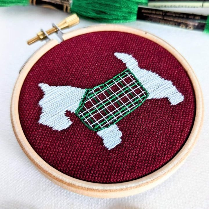 Close up front left angled photo of a white scottie dog with a tartan jacket embroidered on red fabric 