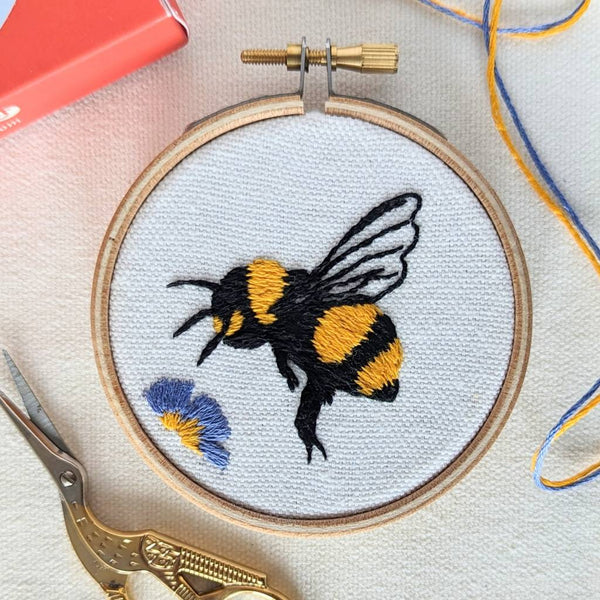 An embroidered honey bee on organic cream fabric, hovering above a King George flower 