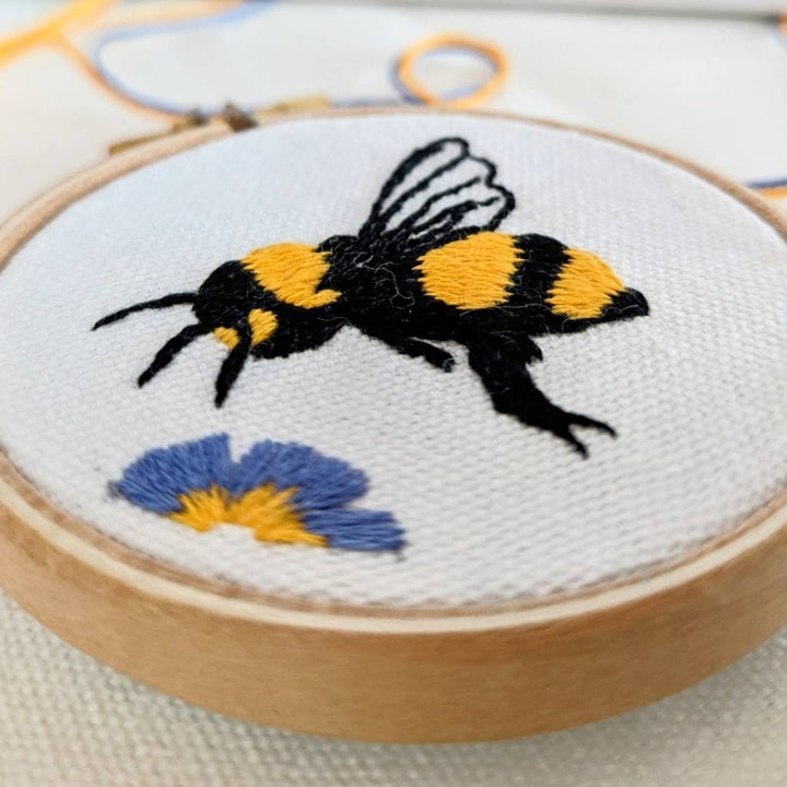 An angled close up photo of an embroidered honey bee on organic cream fabric, hovering above a King George flower 