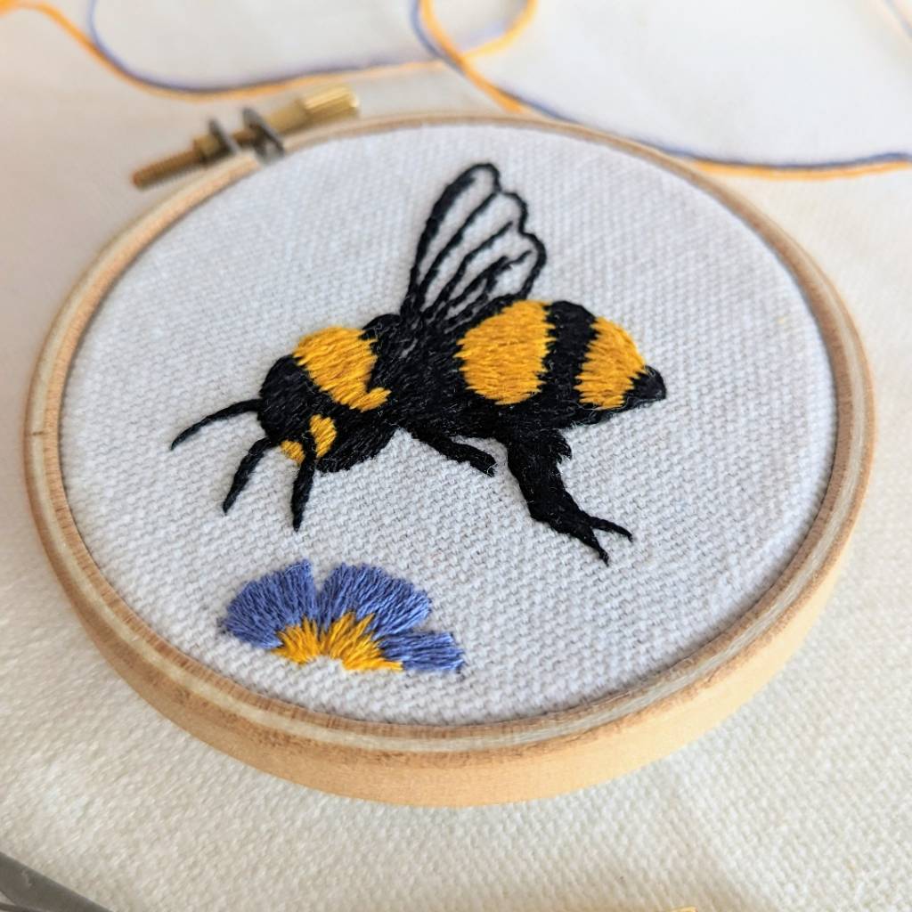 An angled photo of an embroidered honey bee on organic cream fabric, hovering above a King George flower 