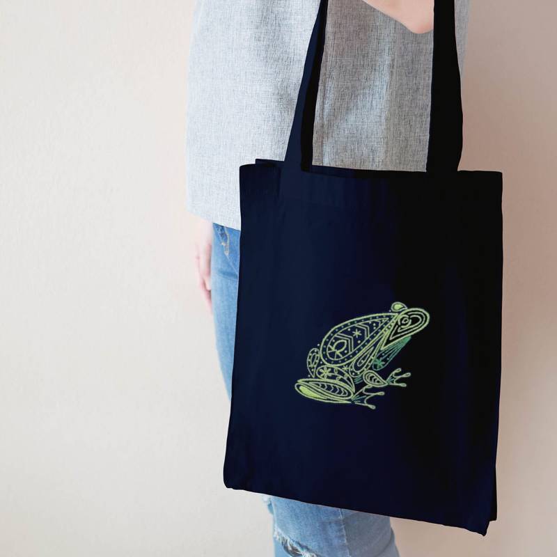 A person holds a navy blue tote bag, embroidered with a green frog design.