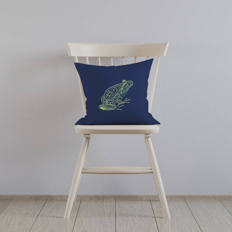 A navy blue cushion embroidered with a green frog design rests upright on a white wooden chair against a white wall.