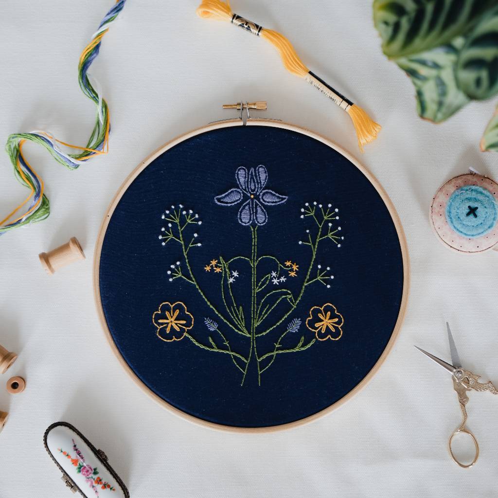 A finished Botanicals embroidery design on navy fabric, with decorative accessories surrounding it. Made using this Botanicals Embroidery kit product.