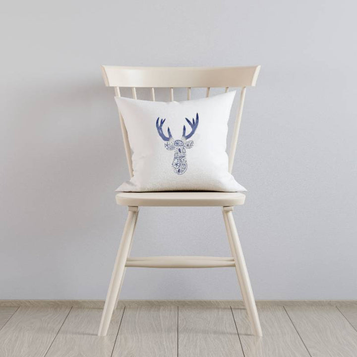 A white cushion sitting on a white wooden chair. The cushion is hand embroidered with a paisley deer pattern in blue and purple thread.