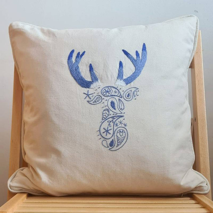 A white cushion sitting on a wooden chair. The cushion is hand embroidered with a paisley deer pattern in blue and purple thread.