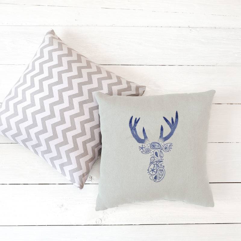 Two cushions lying on white wooden floorboards. The foremost cushion is hand embroidered with a paisley deer pattern in blue and purple thread.