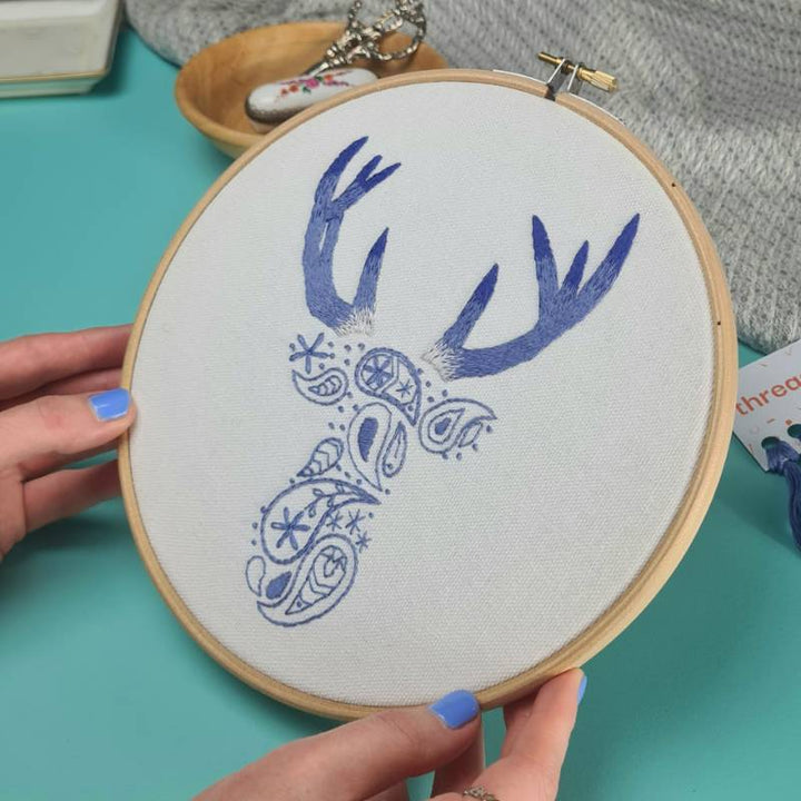 A pair of hands holding a piece of embroidery - a paisley deer design stitched in blue thread on cream fabric.