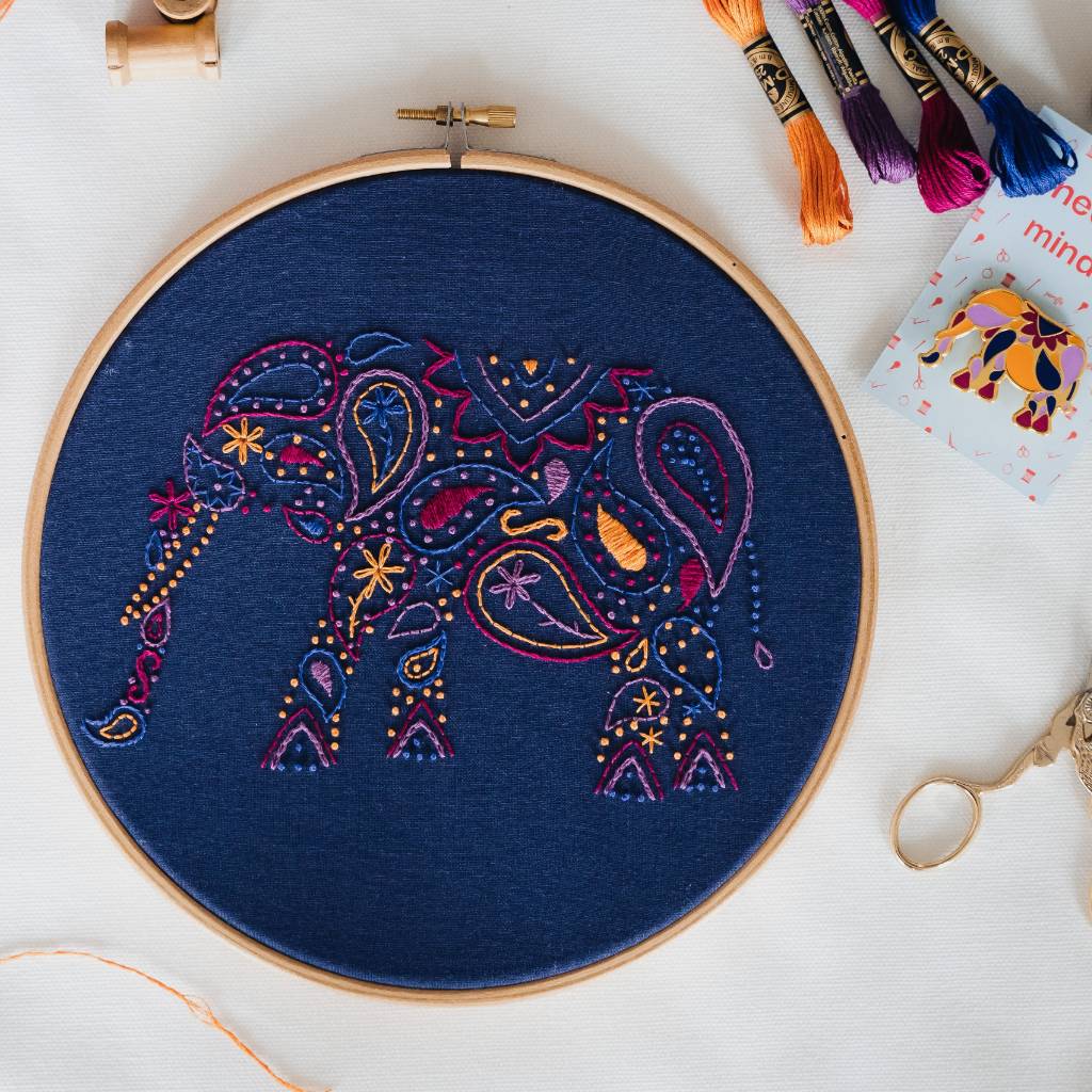 A finished Elephant embroidery design on navy fabric, with scissors, a needleminder and thread surrounding it. Made using this Elephant embroidery kit product.