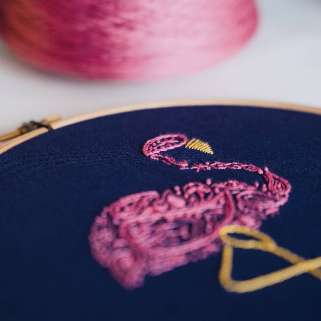 An angled photo of a finished flamingo embroidery design showing the body and legs on navy fabric with pink thread in the background. Made using this flamingo embroidery kit product.