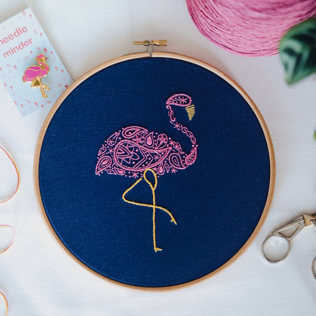 A finished flamingo embroidery design on navy fabric with pink thread and a flamingo needle minder above it and scissors below it. Made using this flamingo embroidery kit product.
