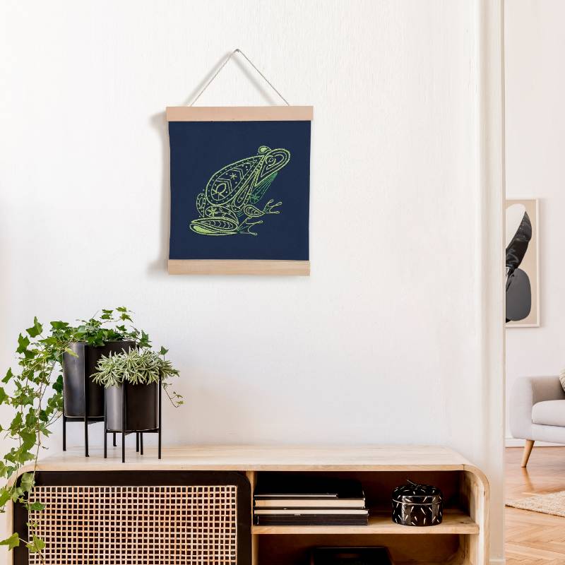 A navy blue square of fabric embroidered with a paisley style frog design hangs between two wooden banner ends, on a white wall, above a wooden cabinet.