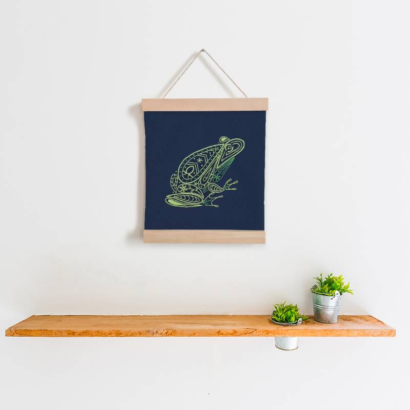 A navy blue square of fabric embroidered with a green frog design hangs between two wooden banner ends, on a white wall, above a wooden shelf.