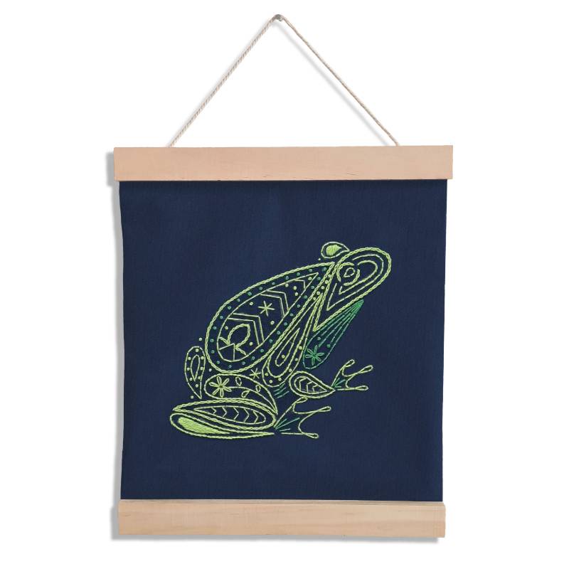 A navy blue piece of fabric hangs between two wooden banner ends against a white backdrop. The fabric is stitched with a paisley style green frog design.