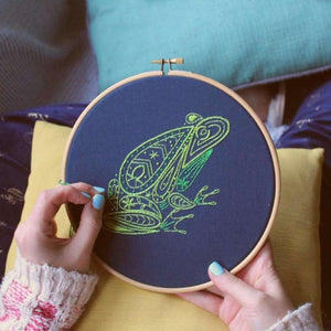 Embroidered frog in two shades of green on a navy fabric in wooden hoop