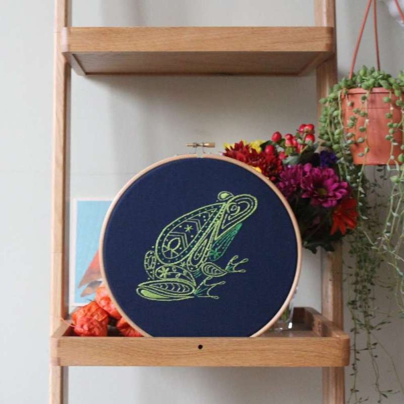 Frog embroidery design in wooden hoop on navy fabric on shelf