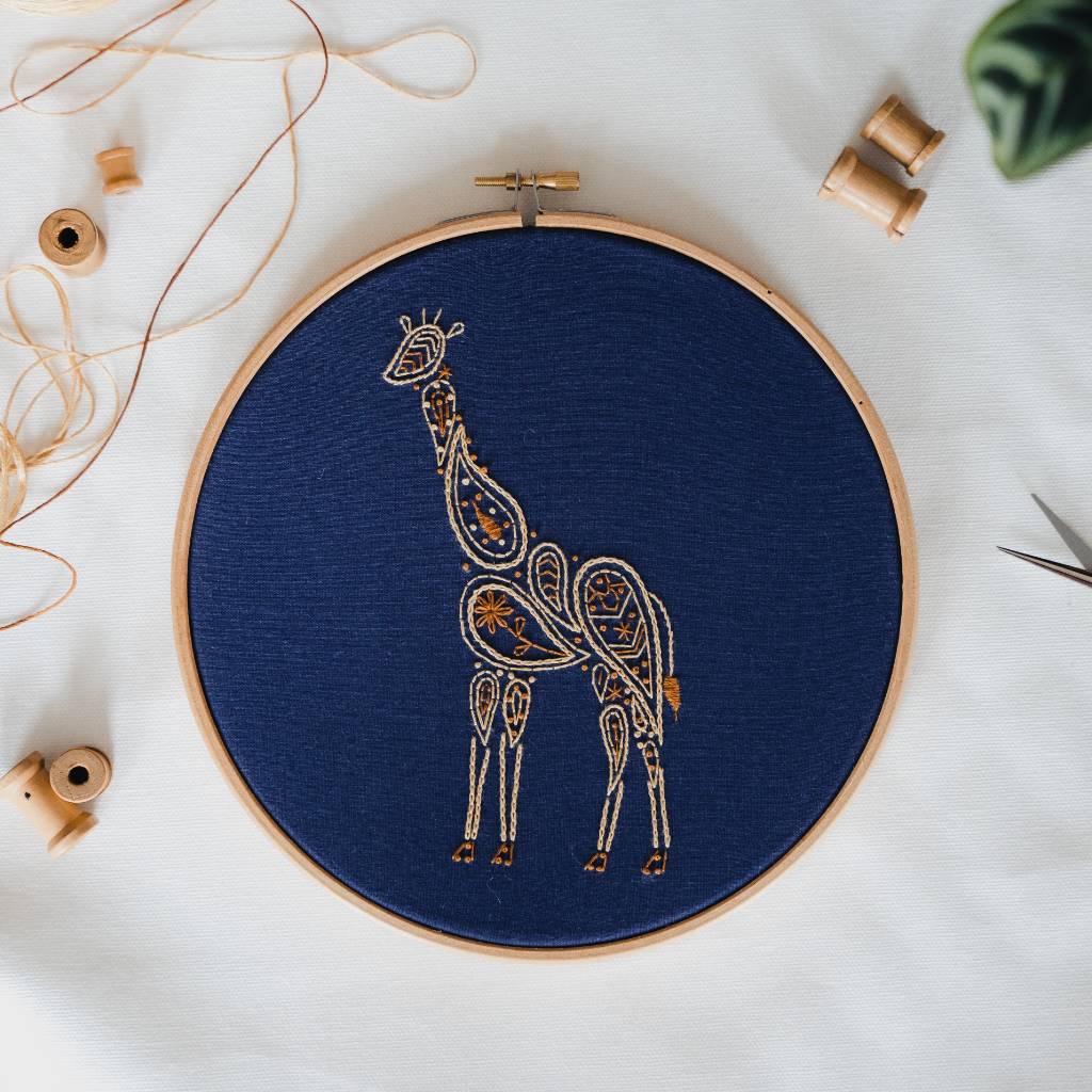 A finished Giraffe embroidery design on navy fabric, with thread and spools surrounding it. Made using this Giraffe embroidery kit product.