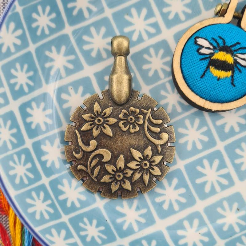A gold embroidery thread cutter in a decorative blue bowl, with a small bee embroidery charm in the top right corner.