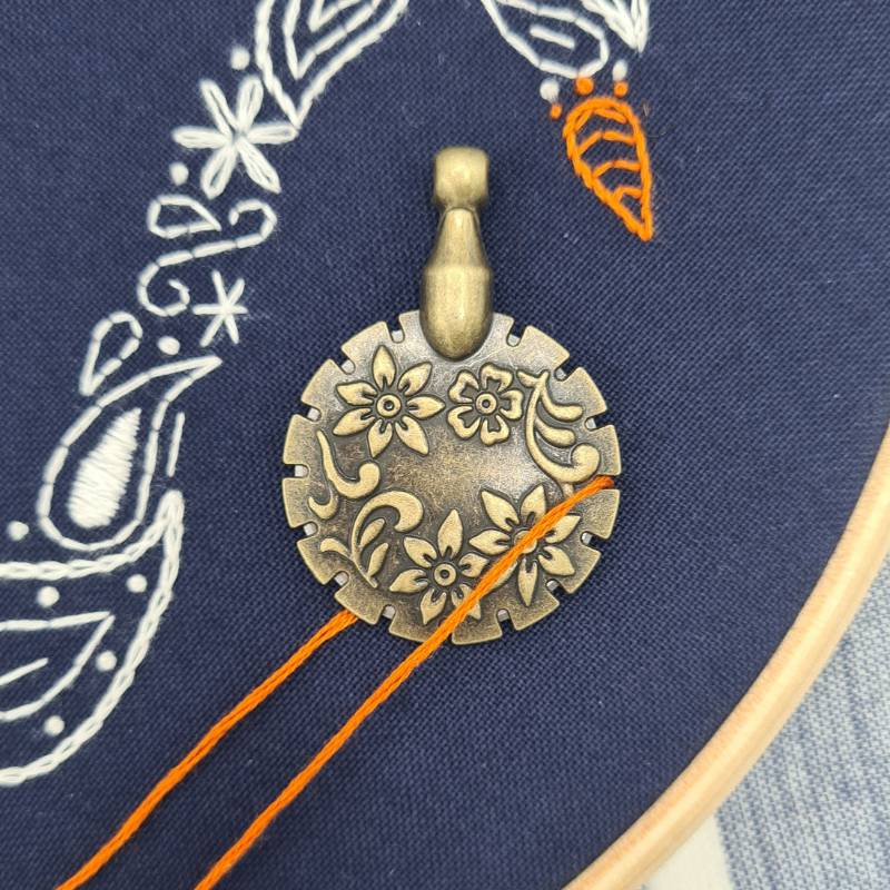 A gold embroidery thread cutter lying on navy fabric, with an orange thread sitting in an outer groove of the cutter.