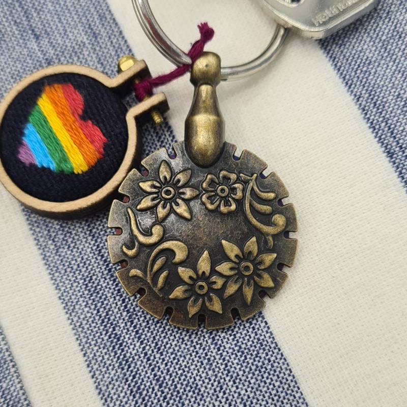 A gold embroidery thread cutter with flower engravings, on a keyring with a rainbow heart embroidery charm.