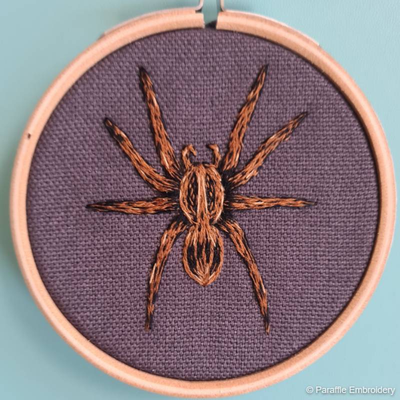 Close up view of an embroidered spider, worked in thread painting style, on grey fabric in a small embroidery hoop