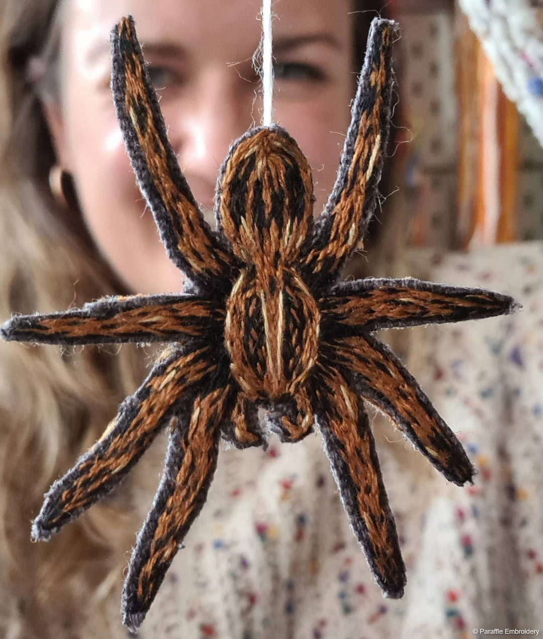 Foreground shows a close up view of an embroidered brown spider, hanging from a white thread. Background shows a partial view of Sammy's face smiling from behind the spider.