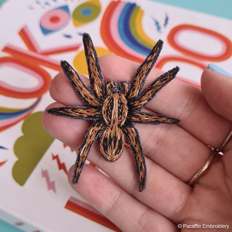 A hand holding an embroidered spider, cut into a patch, stitched in thread painting style