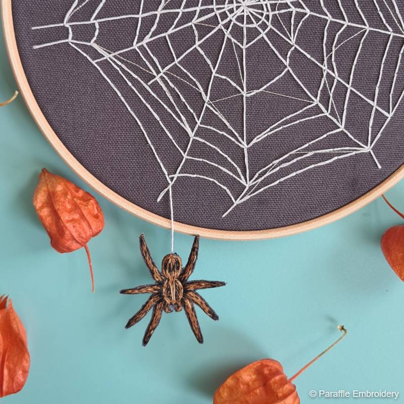 An embroidery hoop stitched with a spider web design; with an embroidered spider hanging from a white thread below it.