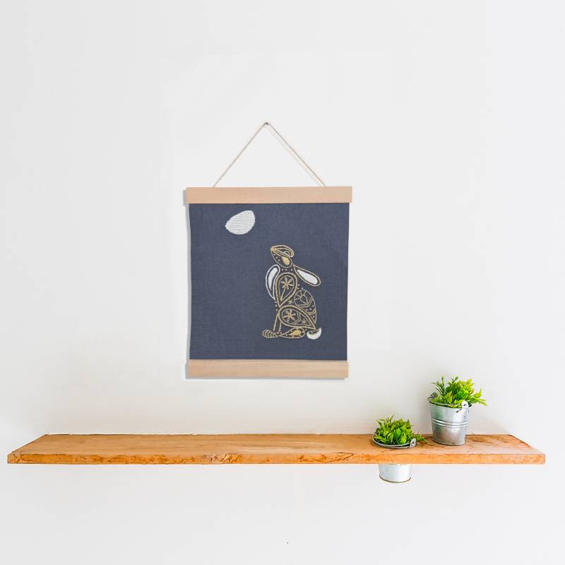 A wooden banner holding a piece of grey fabric embroidered with a moon gazing hare design in a paisley style.