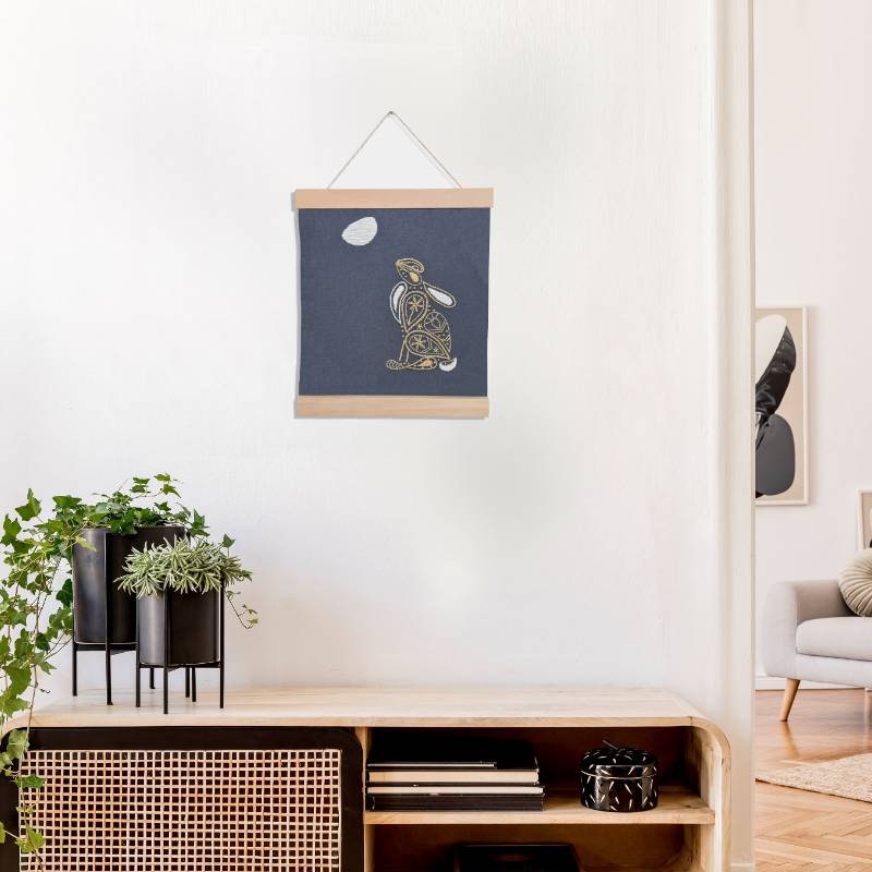 A wooden banner holding a piece of grey fabric embroidered with a moon gazing hare design in a paisley style.