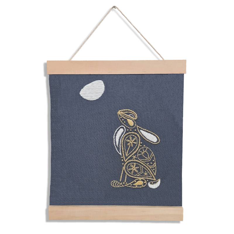 A wooden banner holding a piece of grey fabric embroidered with a moon gazing hare design in a paisley style.