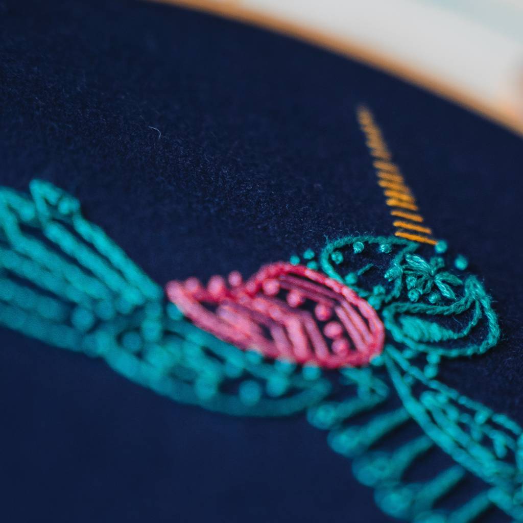 A finished hummingbird embroidery design on navy fabric with thread and decorative accessories surrounding it. Made using this hummingbird embroidery kit product.