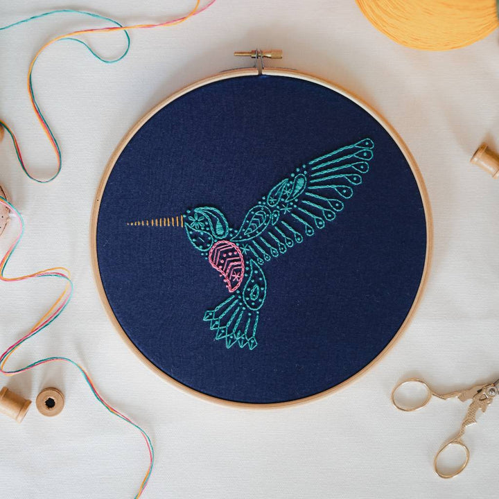 A finished hummingbird embroidery design on navy fabric with thread and decorative accessories surrounding it. Made using this hummingbird embroidery kit product.