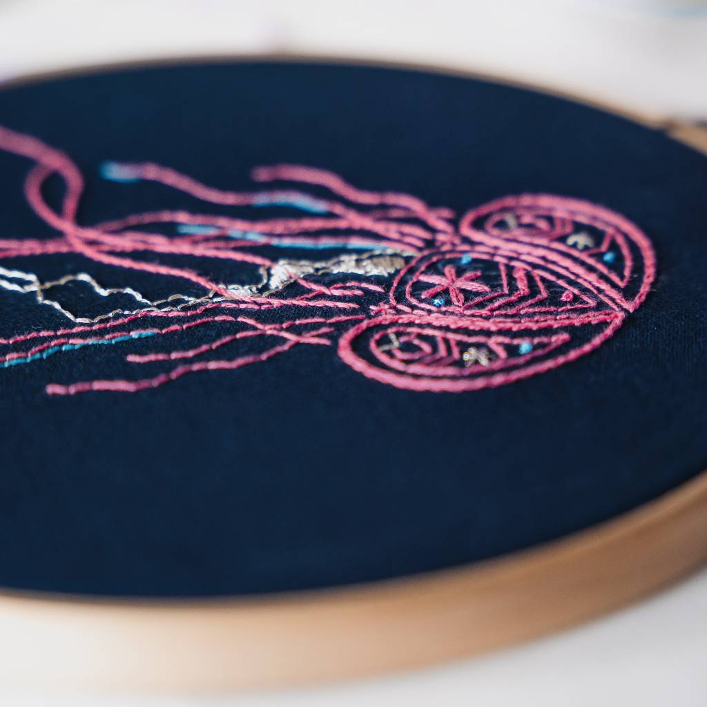 A finished jellyfish embroidery design on navy fabric, with thread and accessories surrounding it. Made using this jellyfish embroidery kit product.