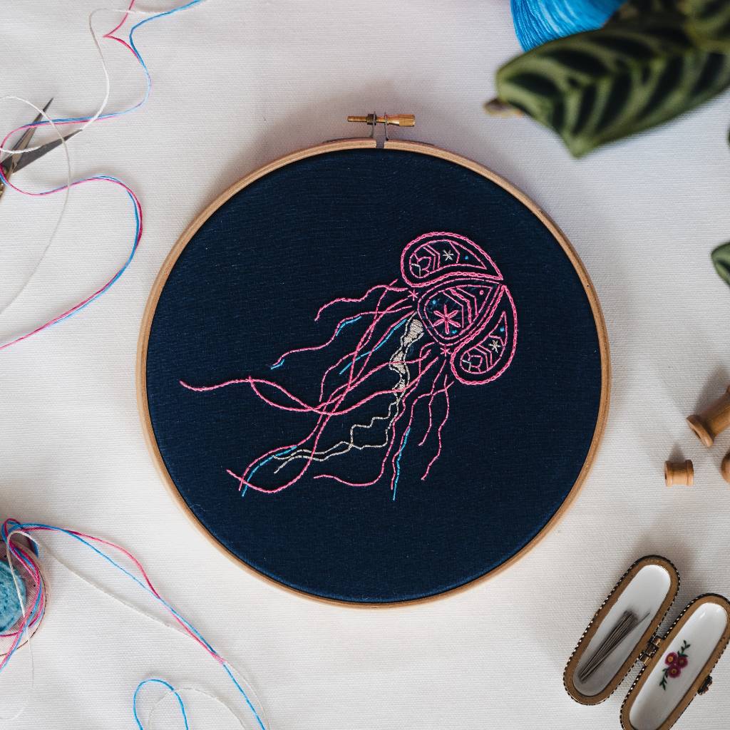 A finished jellyfish embroidery design on navy fabric, with thread and accessories surrounding it. Made using this jellyfish embroidery kit product.