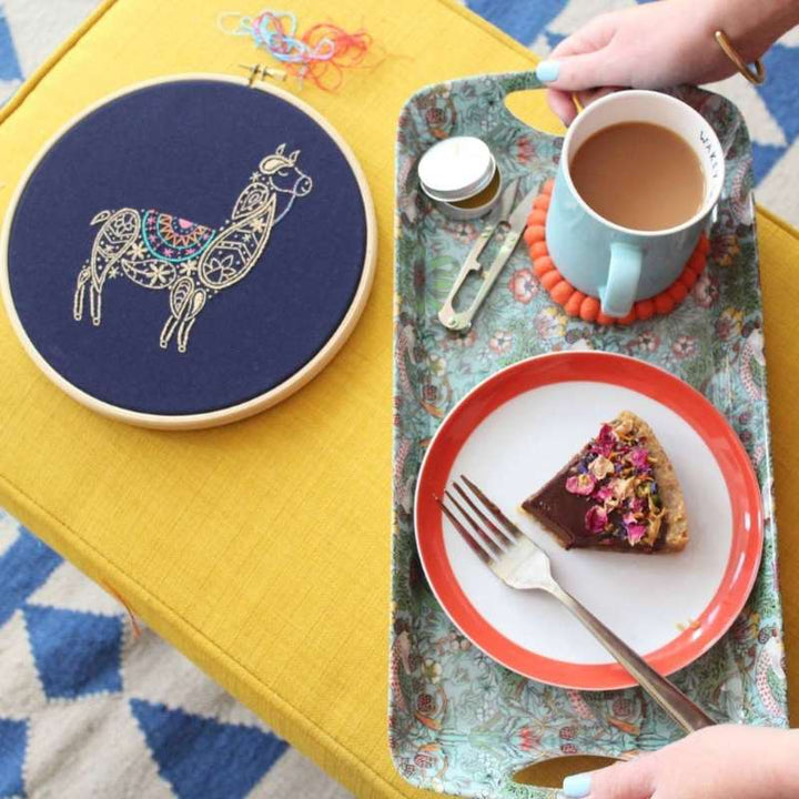 A paisley llama embroidery design in wooden hoop on navy fabric with cake tray next to it