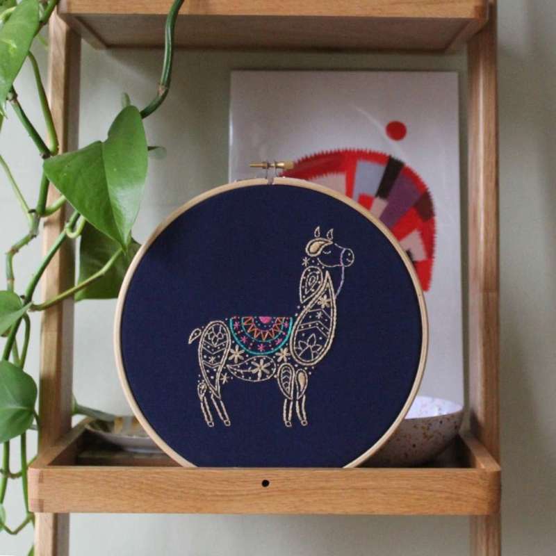 A paisley llama embroidery design in a wooden hoop on navy fabric sat on shelf