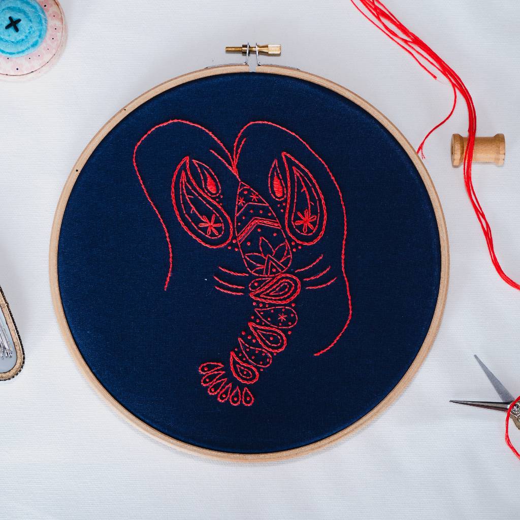 A finished Lobster Embroidery design on Navy fabric, with decorative accessories and red thread surrounding it. Made using this Lobster Embroidery Kit product.