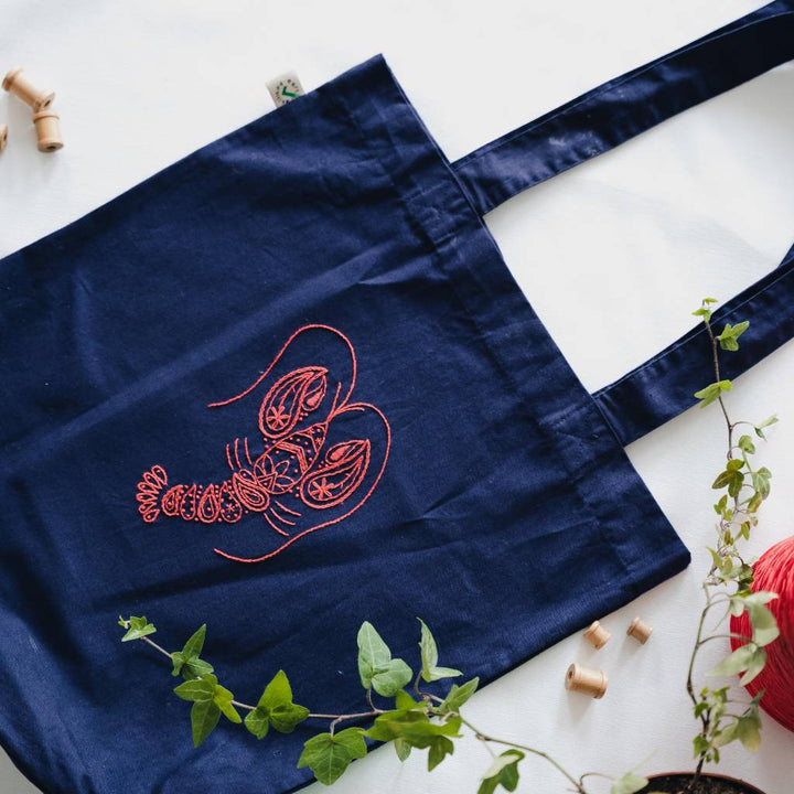 A finished lobster embroidery design embroidered on an organic navy tote bag with thread and a plant in the foreground. Made using this Lobster Tote bag embroidery kit product.