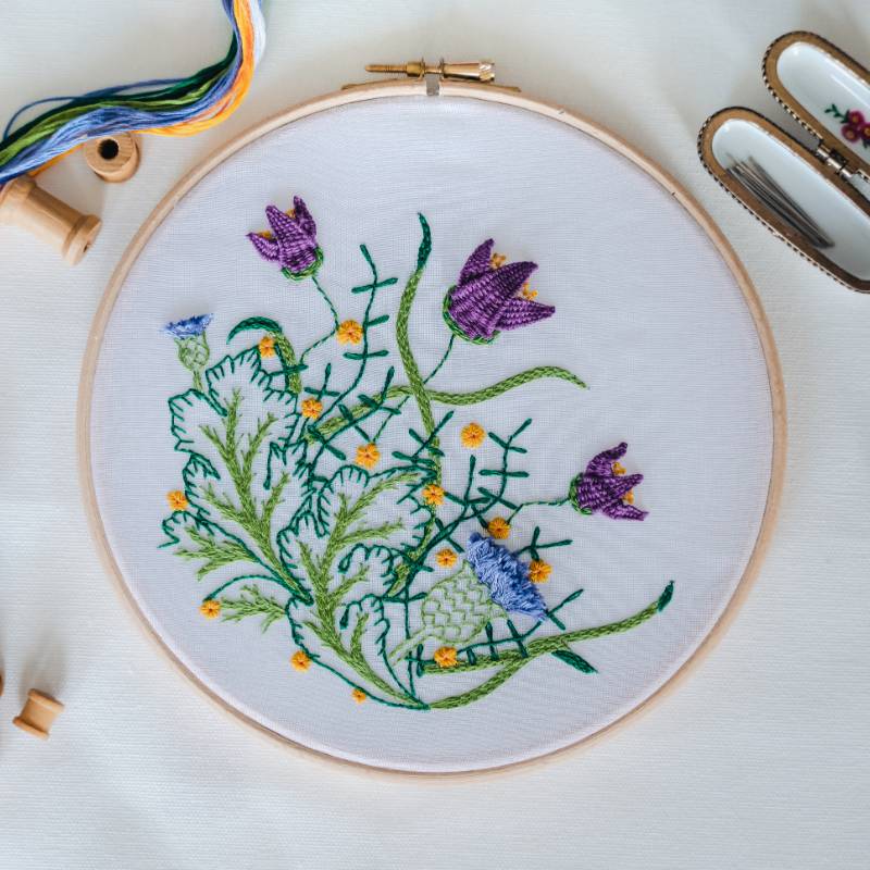 An embroidery hoop containing semi transparent voile fabric, stitched with a William Morris inspired floral design with purple flowers.