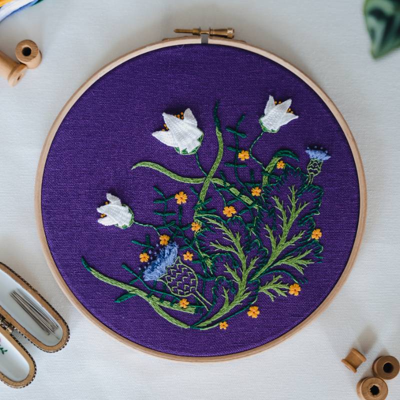 An embroidery hoop containing deep purple fabric, stitched with a William Morris inspired floral design with white flowers.