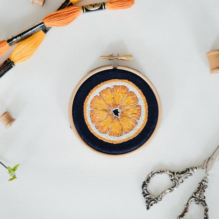 A finished orange slice embroidery design on navy fabric, with scissors below and thread above. 