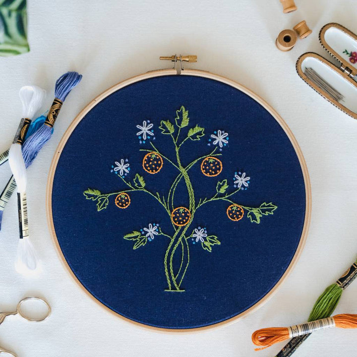 A finished orange tree embroidery design inspired by May Morris on navy fabric with thread and accessories surrounding it. 