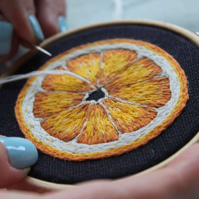 A close up photo of an orange slice embroidered in yellow orange and white thread on navy fabric