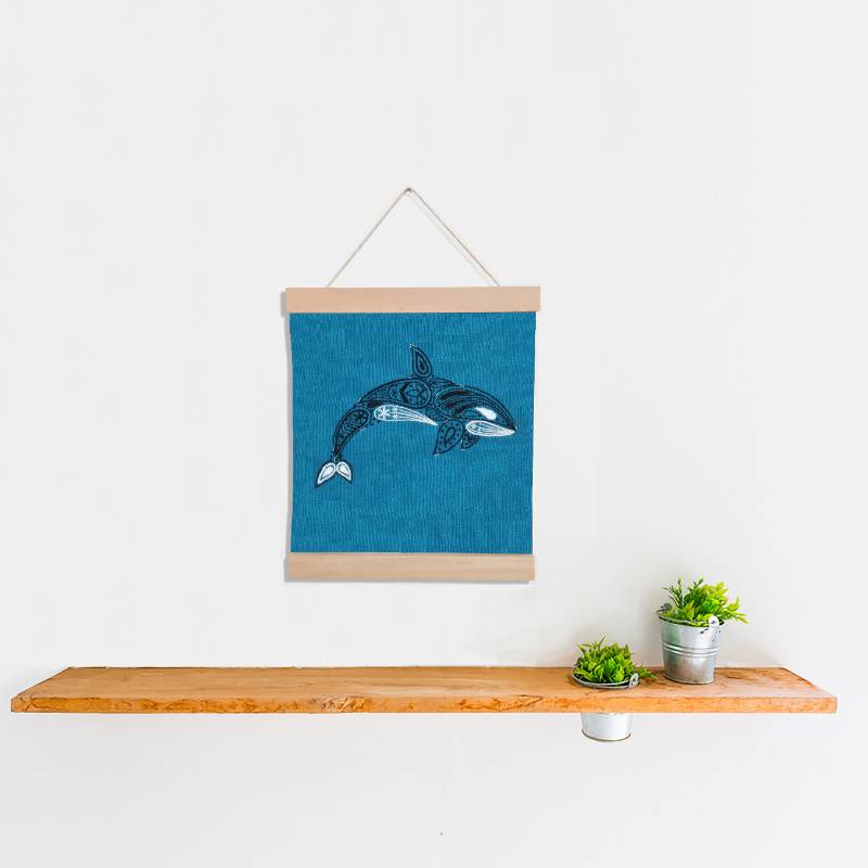 A wooden banner holding a teal square of fabric, set against a white background. The fabric is embroidered with a paisley style orca design.