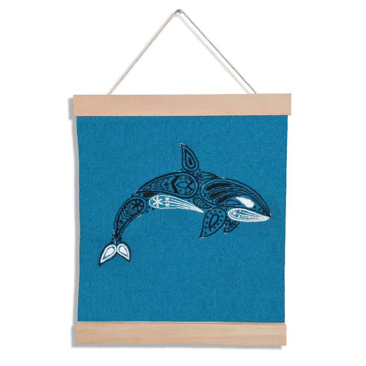 A wooden banner holding a teal square of fabric, set against a white background. The fabric is embroidered with a paisley style orca design.